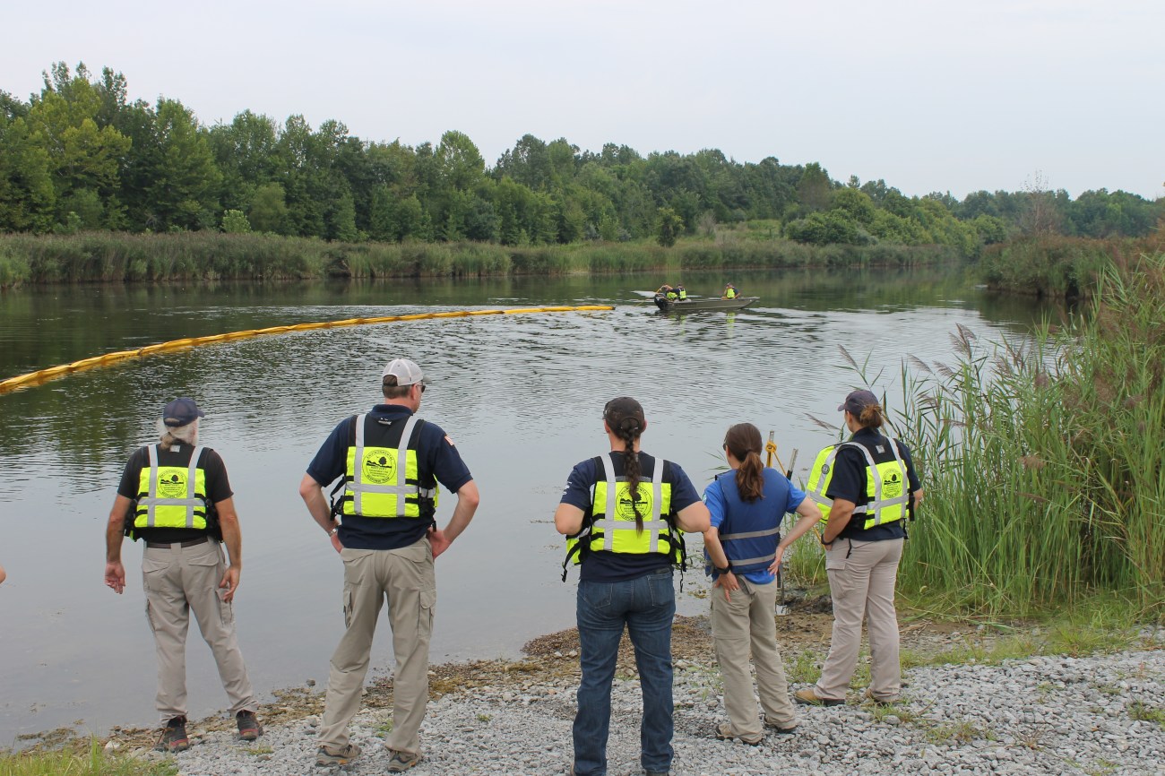 The team practices proper boom deployment and anchoring while being monitored.