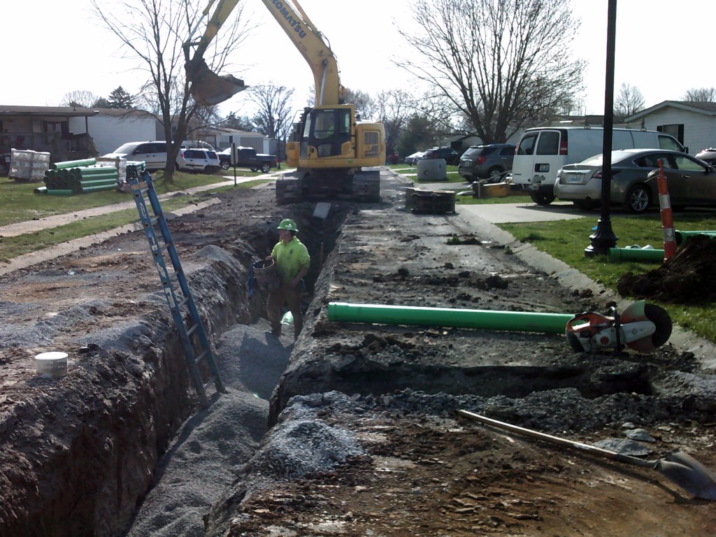 Excavator digs over trench. Man with hard hat and neon yellow shirt holds bucket. 
