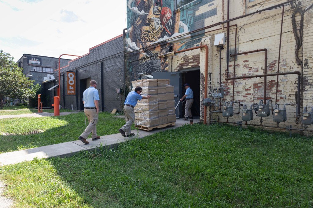 Three men wheel a pallet of boxes into a brick building. Brian Bentley, Jason Craig and Neil Kowalski of the Kentucky Government Recycling Section deliver boxes to FoodChain, a non-profit organization in Lexington on August 15, 2024.