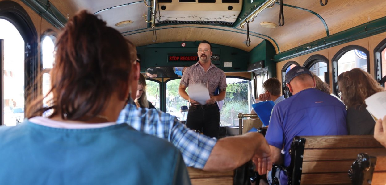 Man in collared shirt addressing passengers on a trolley.