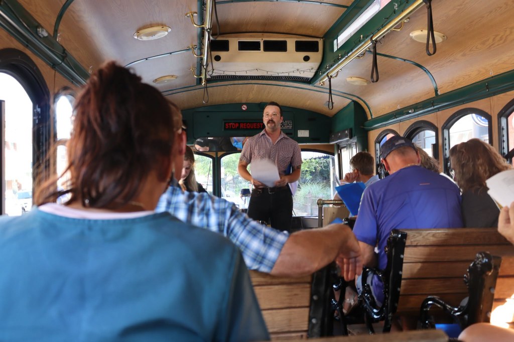 Man in collared shirt addressing passengers on a bus.