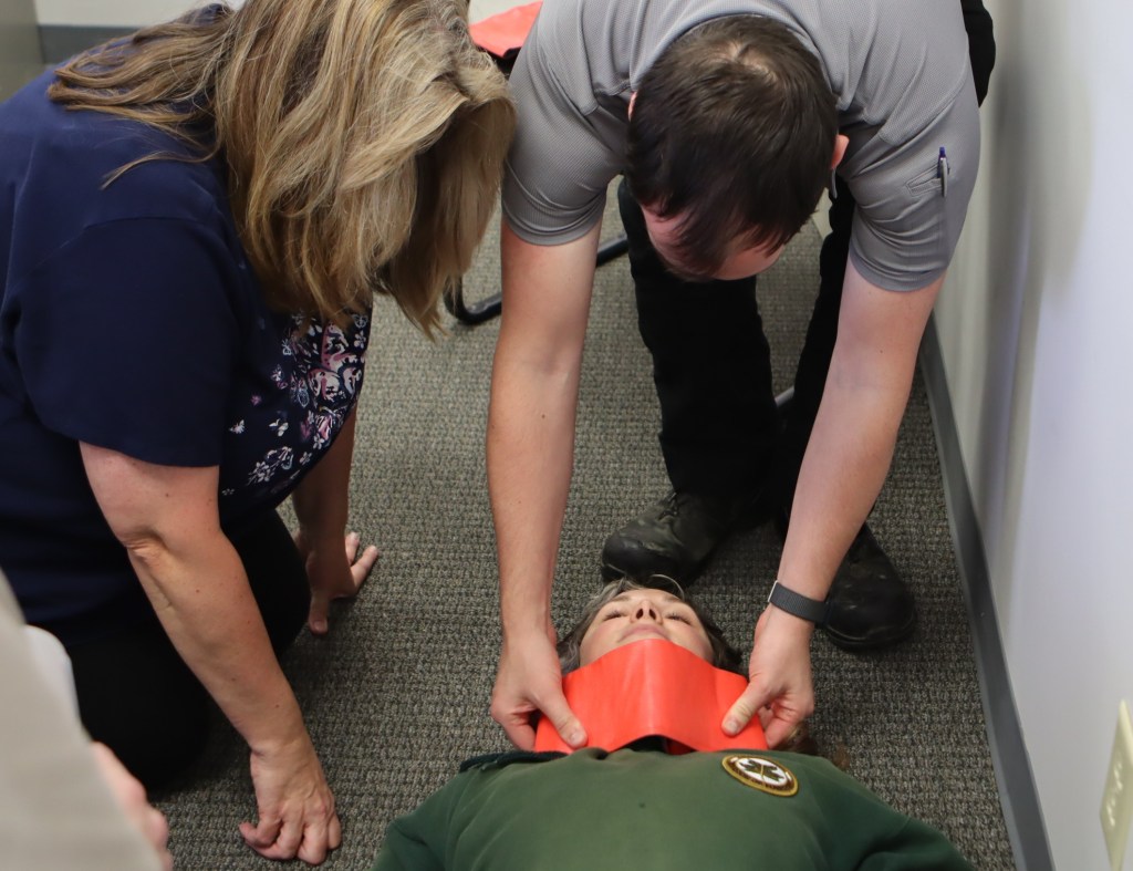 A man demonstrates an improvised neck splint and how to apply in the field on a trainee laying on the floor. 