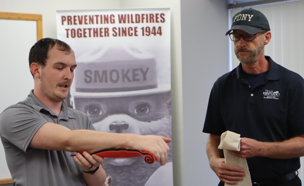 one man is holding an improvised arm splint and another instructor holds a bandage during the demonstration. 